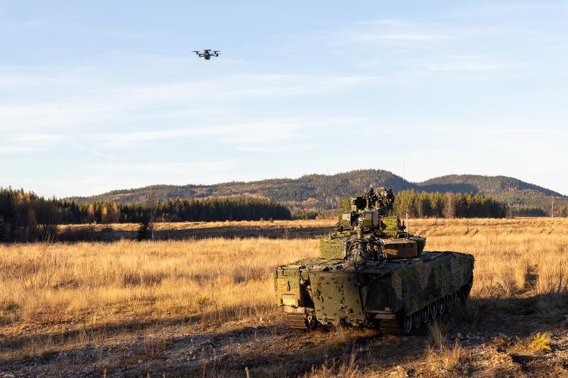 En drone i luften ved en militær beltevogn. Ved en stor slette i høstlig skogslandskap